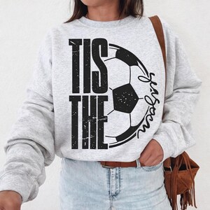 Peut inclure: Un sweat-shirt gris clair avec l'inscription noire "TIS THE SEASON", accompagn&eacute;e d'un ballon de football noir et blanc. Le sweat-shirt a des manches longues et un col rond.