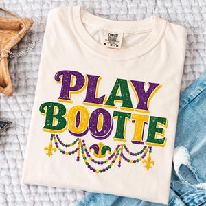May include: Cream-colored t-shirt with the text "PLAY BOOTTE" in purple, green, and yellow. The design includes fleur-de-lis and beaded necklace graphics, evoking a festive theme. The shirt is folded on a denim surface.