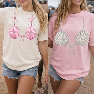 May include: Two short-sleeved t-shirts, one cream and one light pink, each featuring a graphic print of a seashell bikini top. The cream shirt displays a pink seashell design with bows, while the pink shirt shows a silver seashell design with straps.