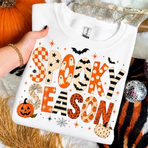 May include: A white crewneck sweatshirt featuring the text "SPOOKY SEASON" in large, patterned letters. The design includes orange and black motifs like ghosts, bats, pumpkins, spiderwebs, and stars, with a Gildan brand label at the neck.