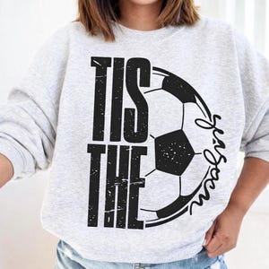 Peut inclure: Un sweat-shirt gris clair avec le texte "Tis the Season" en lettres noires et un graphisme de ballon de football noir et blanc. Le mot "Season" est &eacute;crit en police script.