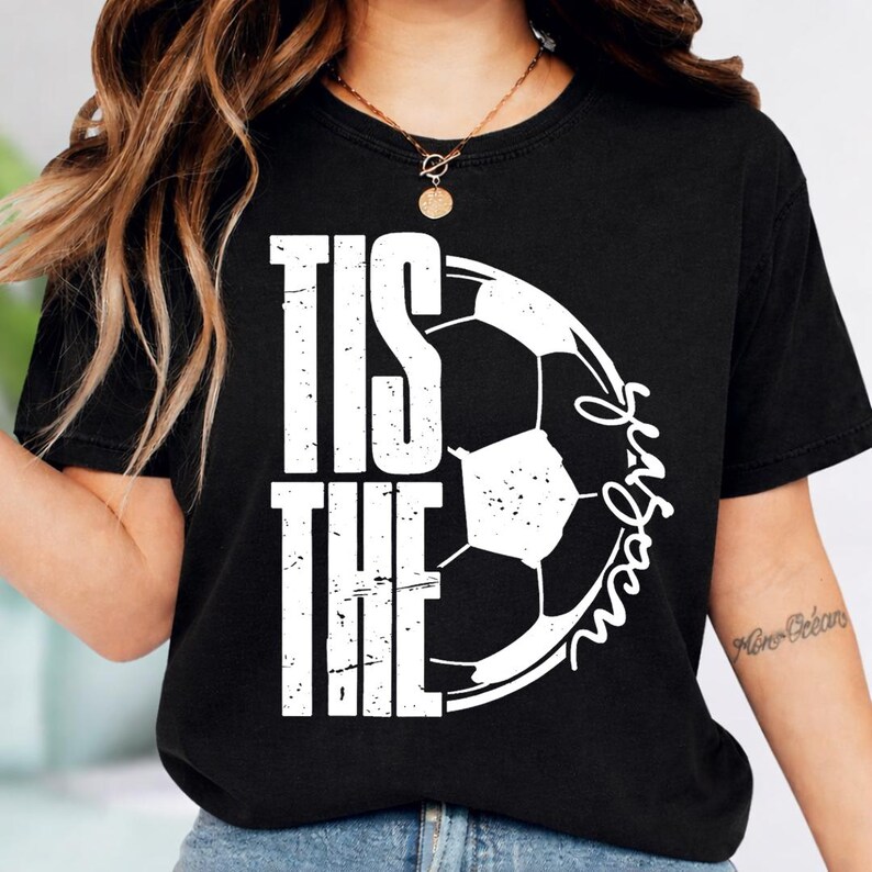 Peut inclure: T-shirt noir avec l'inscription blanche "Tis the Season" en lettres majuscules, accompagn&eacute;e d'un ballon de football blanc. Le mot "Season" est &eacute;crit en cursive autour du ballon.