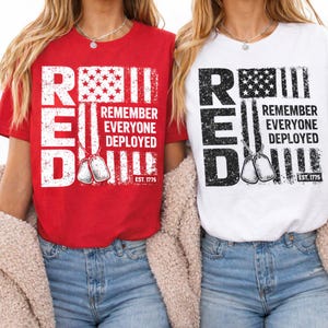 May include: Two t-shirts, one red and one white, each with a patriotic design. The design includes the word "RED" in large block letters, an American flag graphic, the text "REMEMBER EVERYONE DEPLOYED," and dog tag illustrations. The shirts also have "EST. 1775."