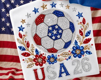 Peut inclure: Un t-shirt blanc orné d'un motif central de ballon de football décoré de strass rouges, blancs et bleus. Le design comprend une disposition circulaire d'étoiles, des accents floraux et le texte USA 26 en strass rouges, blancs et bleus.
