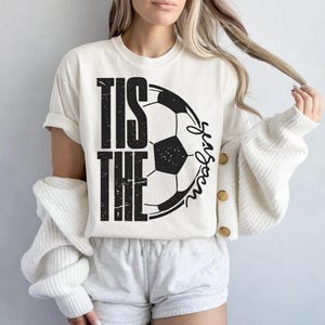 Peut inclure: T-shirt cr&egrave;me avec l'inscription "TIS THE SEASON" en noir, accompagn&eacute;e d'un graphisme de ballon de football. Le mot "Season" est &eacute;crit en script. Le t-shirt est port&eacute; avec un short gris et un cardigan blanc.
