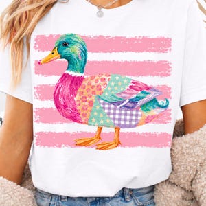 May include: A white short-sleeved t-shirt featuring a colorful mallard duck graphic. The duck has a green head, yellow beak, and a body composed of various patterned patches including stars, gingham, and floral prints, set against horizontal pink stripes.