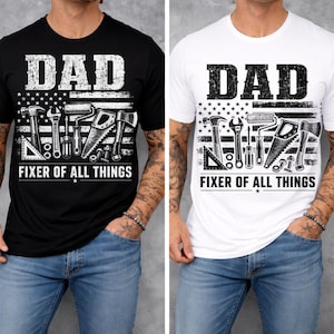 May include: Black and white t-shirts with the word "DAD" above a graphic of tools and an American flag design. The text "FIXER OF ALL THINGS" is below the tools. The shirts are worn by a person.