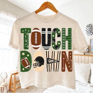 May include: A cream-colored short-sleeve t-shirt featuring the word TOUCHDOWN in large, stylized letters. The design incorporates football-themed graphics, including a brown football, a black helmet, yard lines, and play diagrams.