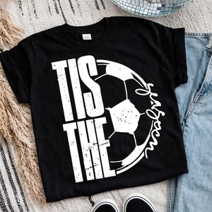Peut inclure: T-shirt noir avec l'inscription blanche "Tis the Season" et un graphisme de ballon de football. Le mot "Season" est &eacute;crit en cursive. Le t-shirt est de style col rond.