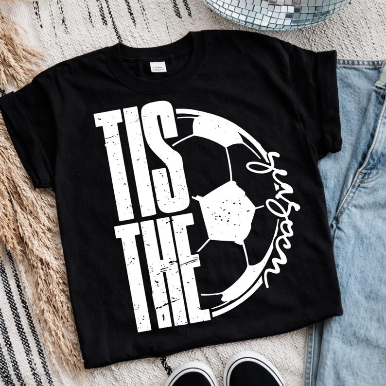 Peut inclure: T-shirt noir avec l'inscription blanche "Tis the Season" et un graphisme de ballon de football. Le mot "Season" est &eacute;crit en cursive. Le t-shirt est de style col rond.