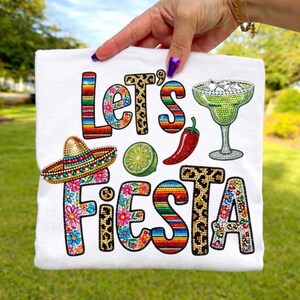 May include: White t-shirt with the words "Let's Fiesta" in colorful, decorative lettering. The design includes a sombrero, margarita glass, lime, and chili pepper, all in a festive style.