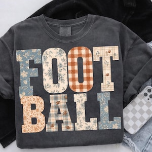 May include: A charcoal gray crewneck t-shirt featuring the word FOOTBALL printed in large block letters. Each letter displays a different pattern, including blue floral, orange gingham, tan plaid, and tan floral, with small football icons.