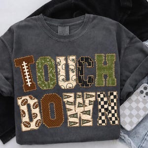 May include: A charcoal gray crew neck t-shirt featuring the word TOUCHDOWN in large block letters. Each letter has a unique pattern including footballs, play diagrams, helmets, mesh textures, game day pennants, and a black and white checkerboard.