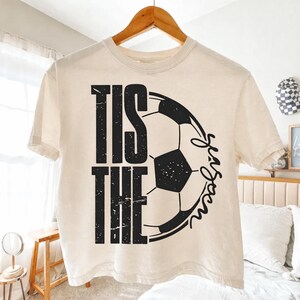 Peut inclure: T-shirt cr&egrave;me avec l'inscription noire "TIS THE SEASON", accompagn&eacute;e d'un graphisme de ballon de football noir et blanc. Le t-shirt est suspendu &agrave; un cintre en bois.