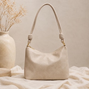 Pu&ograve; includere: Una borsa a spalla color crema con forma curva e cerniera superiore. La borsa ha un manico annodato e finiture color oro. La borsa &egrave; realizzata in un materiale morbido e strutturato.