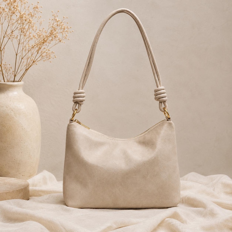 Pu&ograve; includere: Una borsa a spalla color crema con forma curva e cerniera superiore. La borsa ha un manico annodato e finiture color oro. La borsa &egrave; realizzata in un materiale morbido e strutturato.