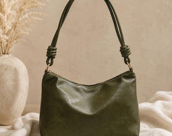 May include: Olive green shoulder bag with a curved shape and a top zipper. The bag has a knotted handle and gold-colored hardware. The bag is sitting on a white cloth.