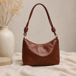 Pu&ograve; includere: Una borsa a tracolla marrone con una forma curva e un manico superiore. La borsa ha una chiusura a cerniera e hardware color oro. Il manico &egrave; attaccato alla borsa con un design annodato. La borsa &egrave; su una superficie color crema.