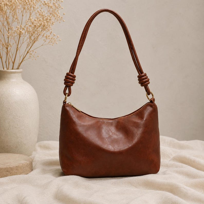 Pu&ograve; includere: Una borsa a tracolla marrone con una forma curva e un manico superiore. La borsa ha una chiusura a cerniera e hardware color oro. Il manico &egrave; attaccato alla borsa con un design annodato. La borsa &egrave; su una superficie color crema.