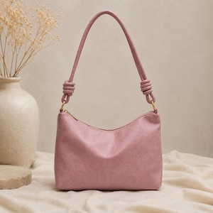 Pu&ograve; includere: Una borsa a spalla rosa cipria con forma curva e cerniera superiore. La borsa ha un manico annodato e finiture color oro. La borsa &egrave; appoggiata su una superficie di colore neutro.