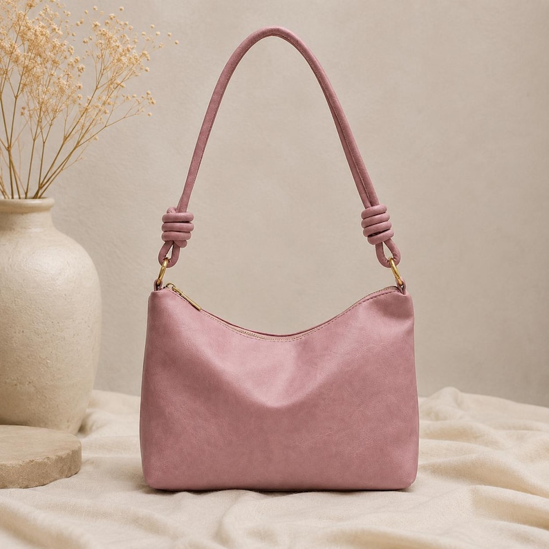 Pu&ograve; includere: Una borsa a spalla rosa cipria con forma curva e cerniera superiore. La borsa ha un manico annodato e finiture color oro. La borsa &egrave; appoggiata su una superficie di colore neutro.
