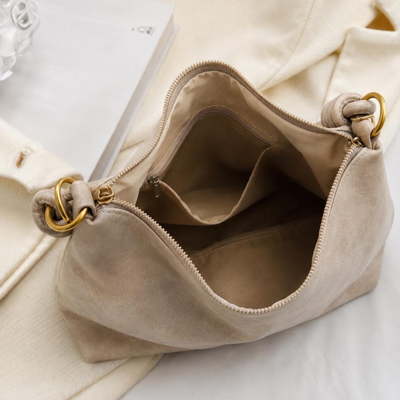 Pu&ograve; includere: Una borsa a mano beige chiaro con chiusura a zip e finiture color oro. L'interno della borsa presenta pi&ugrave; scomparti. La borsa &egrave; realizzata in un materiale morbido simile alla pelle scamosciata e ha una forma arrotondata.