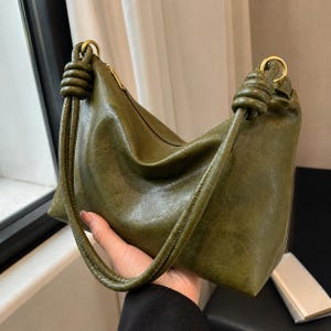 Pu&ograve; includere: Borsa a mano verde oliva con forma curva e chiusura a zip. La borsa presenta manici annodati e dettagli dorati. Il materiale, simile alla pelle, ha un aspetto leggermente invecchiato.