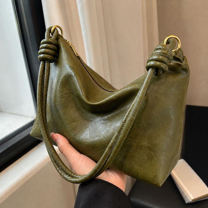 Pu&ograve; includere: Borsa a mano verde oliva con forma curva e chiusura a zip. La borsa presenta manici annodati e dettagli dorati. Il materiale, simile alla pelle, ha un aspetto leggermente invecchiato.