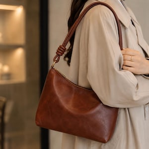 Pu&ograve; includere: Una borsa a tracolla in pelle marrone con forma curva e chiusura a zip. La borsa ha una tracolla marrone abbinata e finiture color oro. La borsa &egrave; indossata da una persona che indossa un cappotto beige.