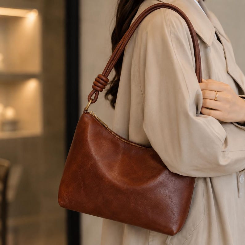Pu&ograve; includere: Una borsa a tracolla in pelle marrone con forma curva e chiusura a zip. La borsa ha una tracolla marrone abbinata e finiture color oro. La borsa &egrave; indossata da una persona che indossa un cappotto beige.