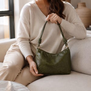 Pu&ograve; includere: Borsa a spalla verde oliva con dettagli a nodo sulle cinghie. La borsa ha una forma curva e una cerniera dorata. La borsa &egrave; realizzata in un materiale strutturato. Una persona indossa un maglione color crema.