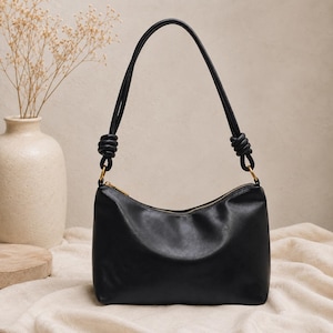 Pu&ograve; includere: Borsa a tracolla in pelle nera con chiusura a zip curva e design con manico annodato. La borsa ha una cerniera dorata e una forma semplice ed elegante. Un vaso con fiori secchi &egrave; sullo sfondo.