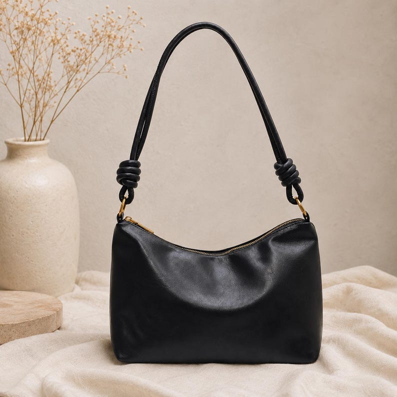 Pu&ograve; includere: Borsa a tracolla in pelle nera con chiusura a zip curva e design con manico annodato. La borsa ha una cerniera dorata e una forma semplice ed elegante. Un vaso con fiori secchi &egrave; sullo sfondo.