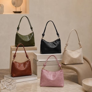 Pu&ograve; includere: Cinque borse a tracolla in pelle di vari colori: verde oliva, nero, marrone, rosa e beige. Ogni borsa ha un manico annodato e una forma curva. Le borse sono esposte su uno sfondo neutro, alcune su scatole impilate.