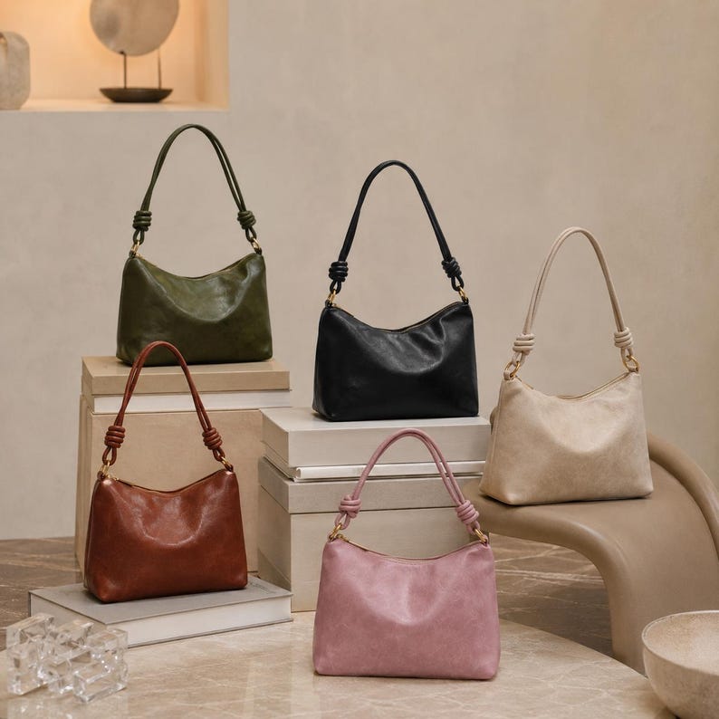 Pu&ograve; includere: Cinque borse a tracolla in pelle di vari colori: verde oliva, nero, marrone, rosa e beige. Ogni borsa ha un manico annodato e una forma curva. Le borse sono esposte su uno sfondo neutro, alcune su scatole impilate.