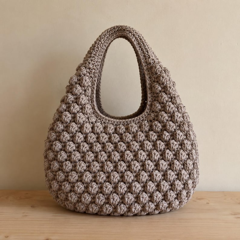May include: A beige crochet handbag featuring a prominent bobble stitch texture throughout the exterior. The bag has a rounded, teardrop-like silhouette with a single integrated handle opening at the top, showcasing a consistent, dense, and uniform pattern.