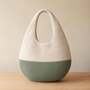 May include: A teardrop-shaped handbag crafted from crocheted yarn, featuring a two-tone color scheme with a cream-colored upper section and a sage green lower section. The bag has a single integrated handle at the top and a textured surface.