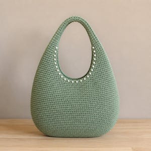 May include: A sage green crochet handbag with a teardrop shape and a single integrated handle. The inner edge of the handle is decorated with a row of small, round, white faux pearls that follow the curve of the opening of the bag.