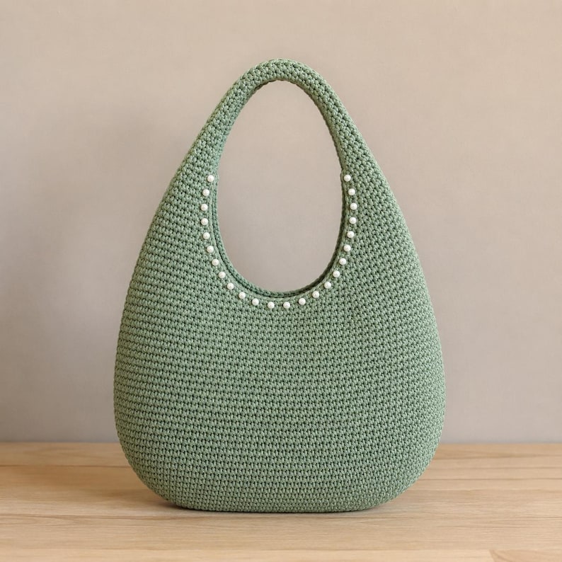 May include: A sage green crochet handbag with a teardrop shape and a single integrated handle. The inner edge of the handle is decorated with a row of small, round, white faux pearls that follow the curve of the opening of the bag.