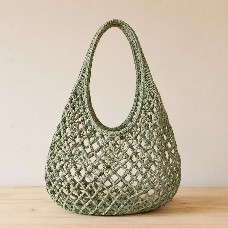 May include: A sage green crochet tote bag featuring an open diamond-shaped mesh pattern throughout the body. The bag has a single integrated handle at the top, constructed with a solid, dense crochet border that transitions into the open weave.