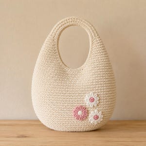 May include: A cream-colored crochet handbag with a rounded, teardrop-shaped silhouette and an integrated handle. The bag features three small, flat, stitched floral appliqu&eacute;s on the lower right side, each with white petals and a pink center.
