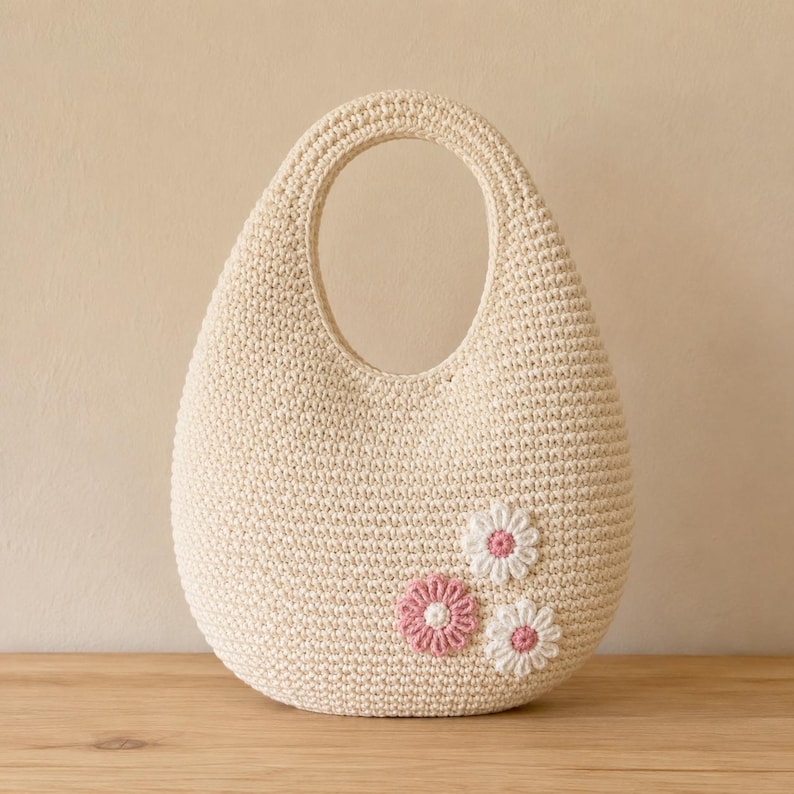 May include: A cream-colored crochet handbag with a rounded, teardrop-shaped silhouette and an integrated handle. The bag features three small, flat, stitched floral appliqu&eacute;s on the lower right side, each with white petals and a pink center.