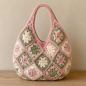 May include: A crochet handbag constructed from multiple square motifs in shades of soft pink, sage green, and cream. The bag features a rounded, teardrop-like silhouette with a single integrated handle, showcasing a repeating floral pattern.
