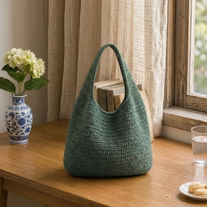 May include: A sage green hobo-style handbag crafted with a textured, woven knit pattern. The bag features a single integrated handle and a spacious, rounded silhouette, showcasing a consistent, dense stitch throughout its entire exterior surface.