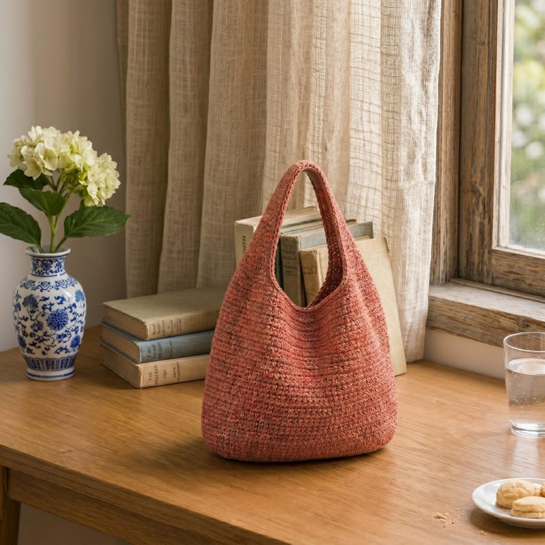 May include: A terracotta-colored, woven hobo-style handbag with a single integrated handle. The bag features a dense, textured knit construction and a rounded base, standing upright on a wooden surface with a visible weave pattern throughout.