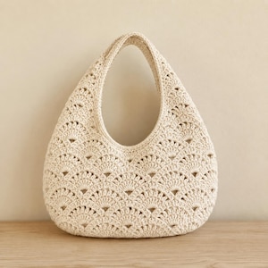 May include: A cream-colored hobo-style handbag crafted with a detailed crochet technique. The bag features a repeating fan-shaped stitch pattern throughout its exterior, a single integrated handle, and a rounded, teardrop-like silhouette.
