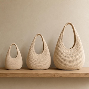 May include: Three beige crochet bags of varying sizes arranged in a row. Each bag features a rounded, teardrop-shaped body with a single integrated handle at the top, crafted from a dense, uniform stitch pattern in a neutral light tan color.