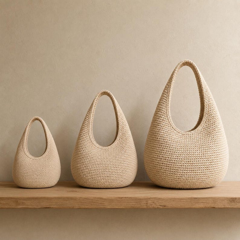 May include: Three beige crochet bags of varying sizes arranged in a row. Each bag features a rounded, teardrop-shaped body with a single integrated handle at the top, crafted from a dense, uniform stitch pattern in a neutral light tan color.