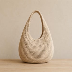 May include: A beige hobo-style handbag crafted with a dense, uniform crochet stitch pattern. The bag features a rounded, teardrop-shaped silhouette with a single integrated handle that forms a large, open oval opening at the top of the structure.