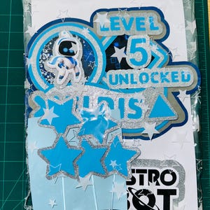 May include: A selection of Astro Bot themed cake decorations in blue and silver. The toppers feature the text "LEVEL 5 UNLOCKED" and "ASTRO BOT", alongside star and character cutouts. The items are packaged in a clear plastic bag.